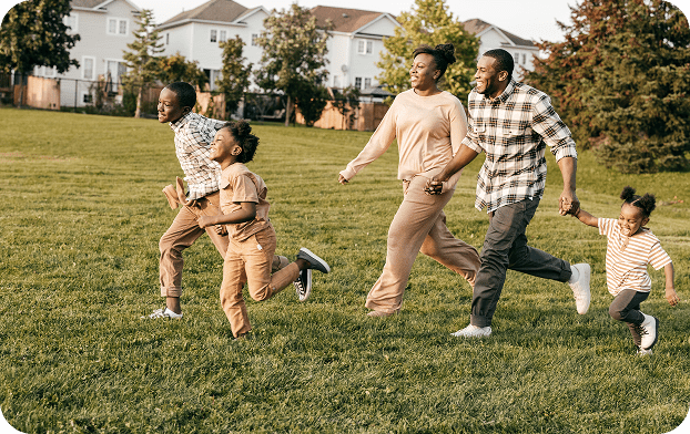 A family of four running and playing on grass in a sunny park.