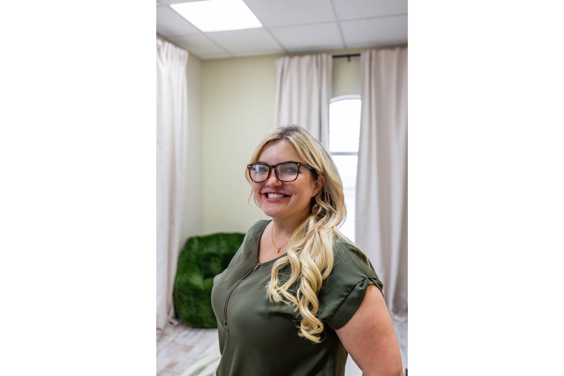 Smiling woman with long blonde hair and glasses in a bright room.